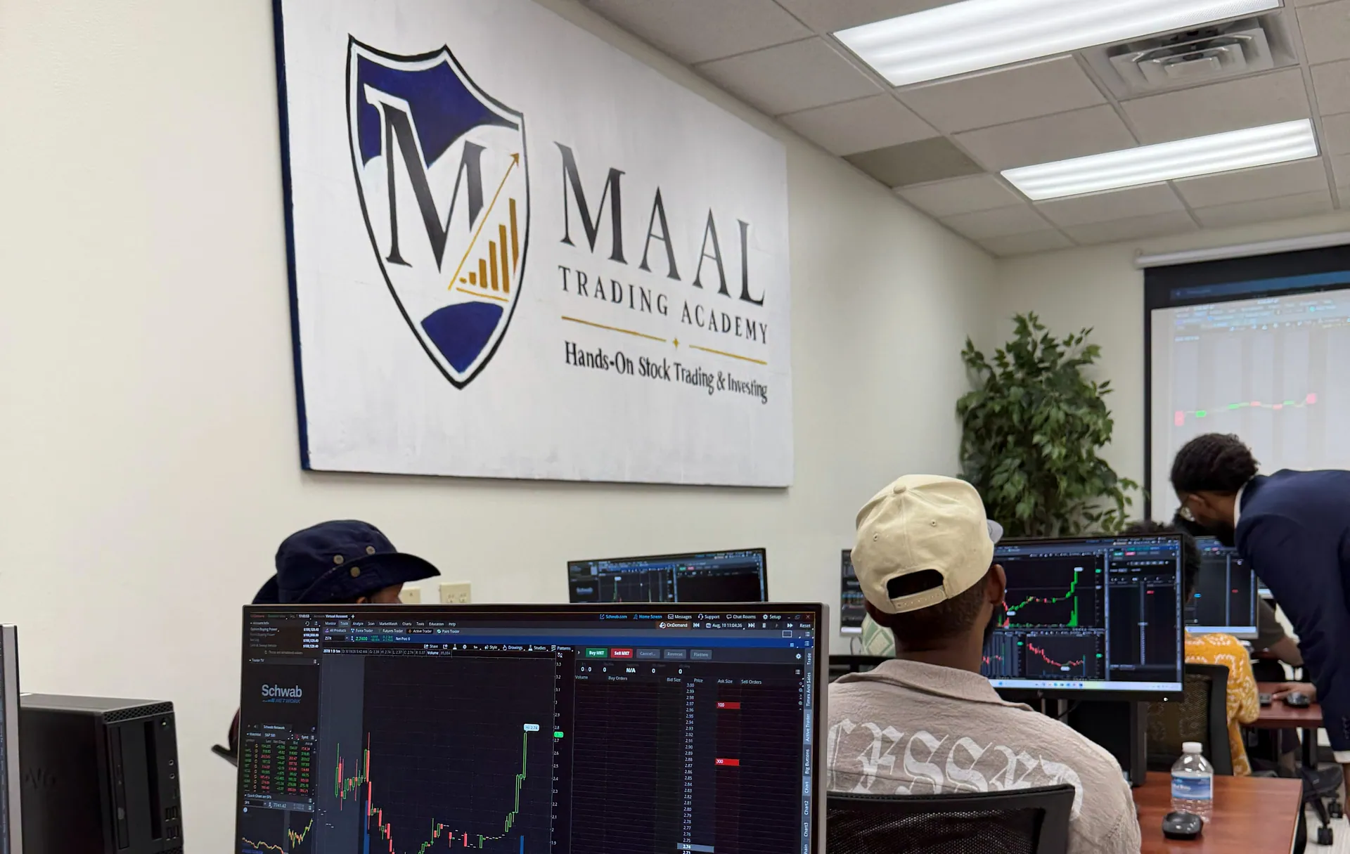The MAAL TRADING ACADEMY wall sign above a class at their stations in Minneapolis, a live chart on the nearest screen.