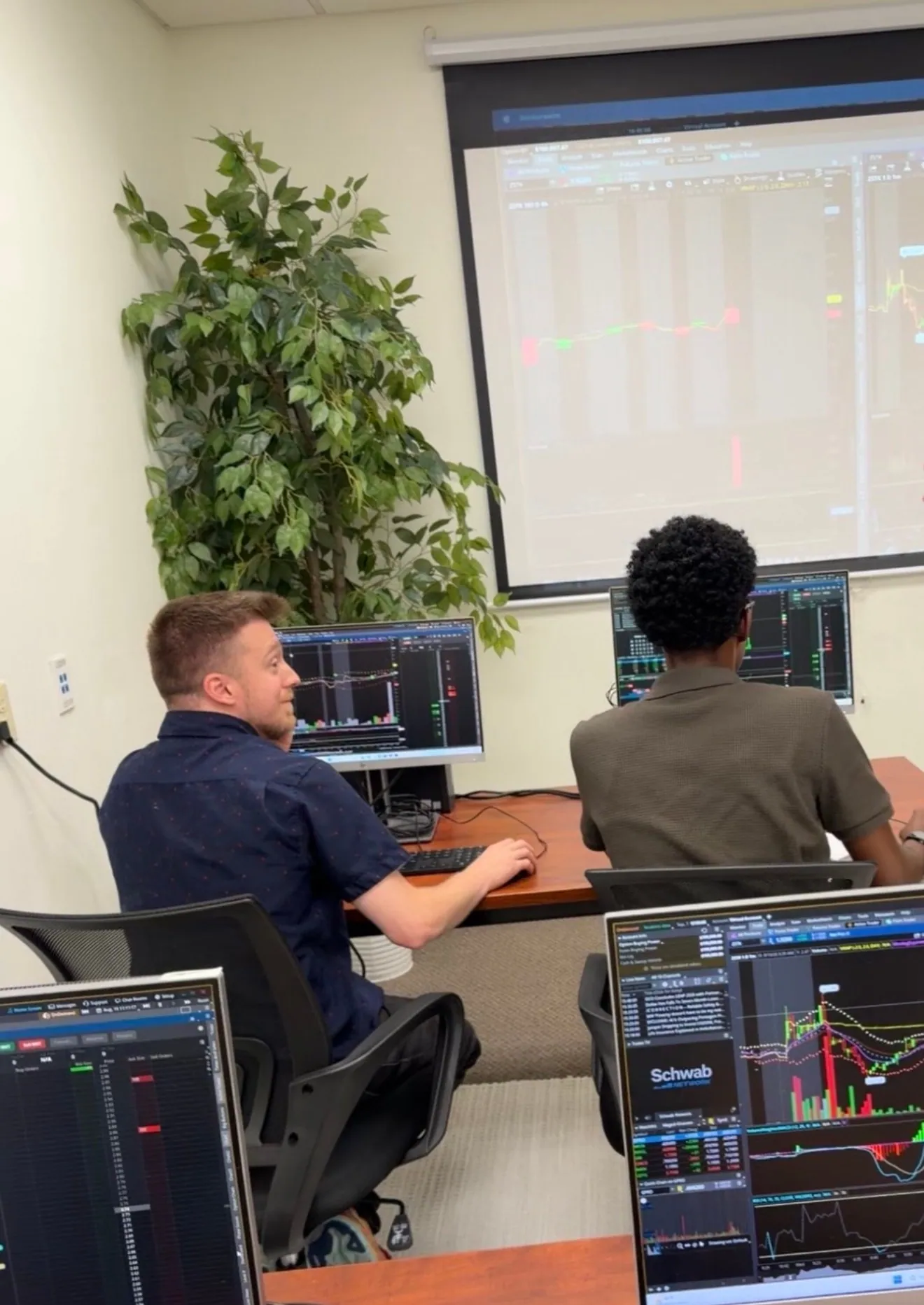 Two beginner students at their Schwab trading stations at MAAL TRADING ACADEMY, a live chart on the big screen behind them.